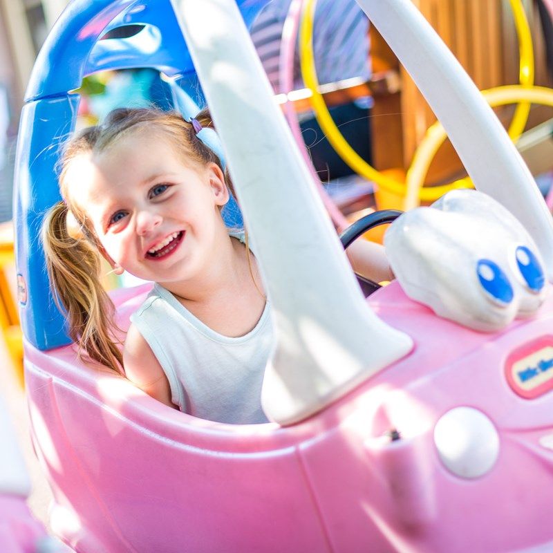 A toddler grinning from the seat of a pink toy car